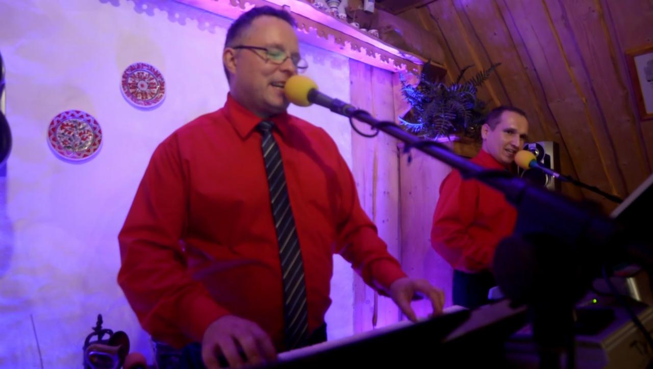 Zespół muzyczny w czerwonych koszulach grający na keyboardzie i śpiewający do mikrofonu na scenie z drewnianym sufitem i dekoracyjnymi talerzami na ścianie.