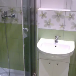 Umywalka z szafką, toaleta i kabina prysznicowa w łazience wyłożonej zielono-białymi płytkami z motywem kwiatowym.