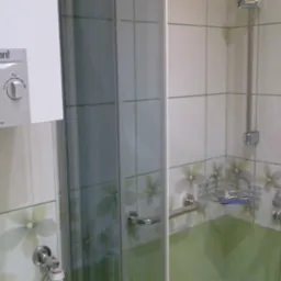 Biały piec gazowy marki Vaillant zamontowany nad umywalką w łazience z kabiną prysznicową, wykończoną kafelkami w biało-zielone kwiaty.