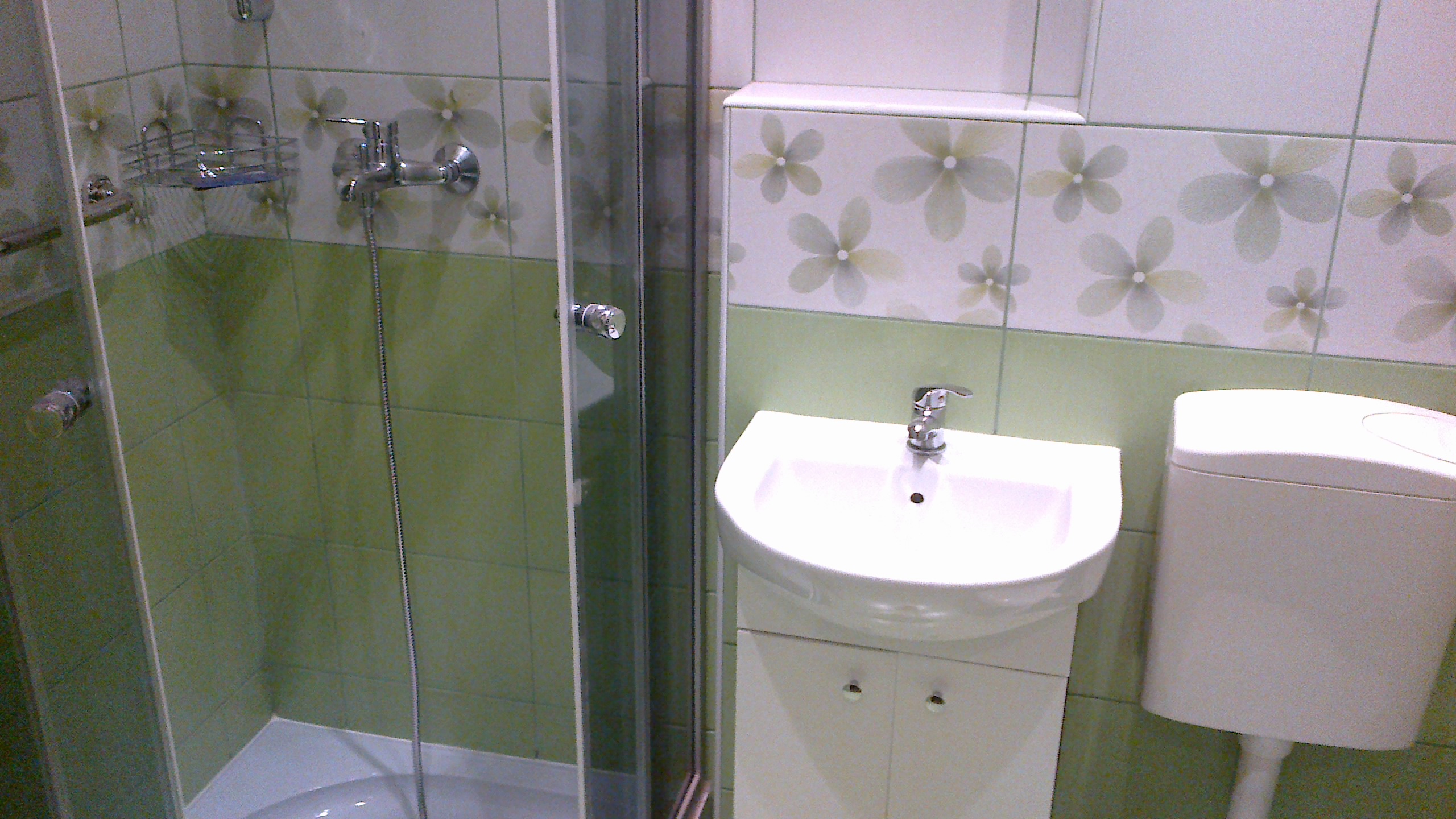 Umywalka z szafką, toaleta i kabina prysznicowa w łazience wyłożonej zielono-białymi płytkami z motywem kwiatowym.