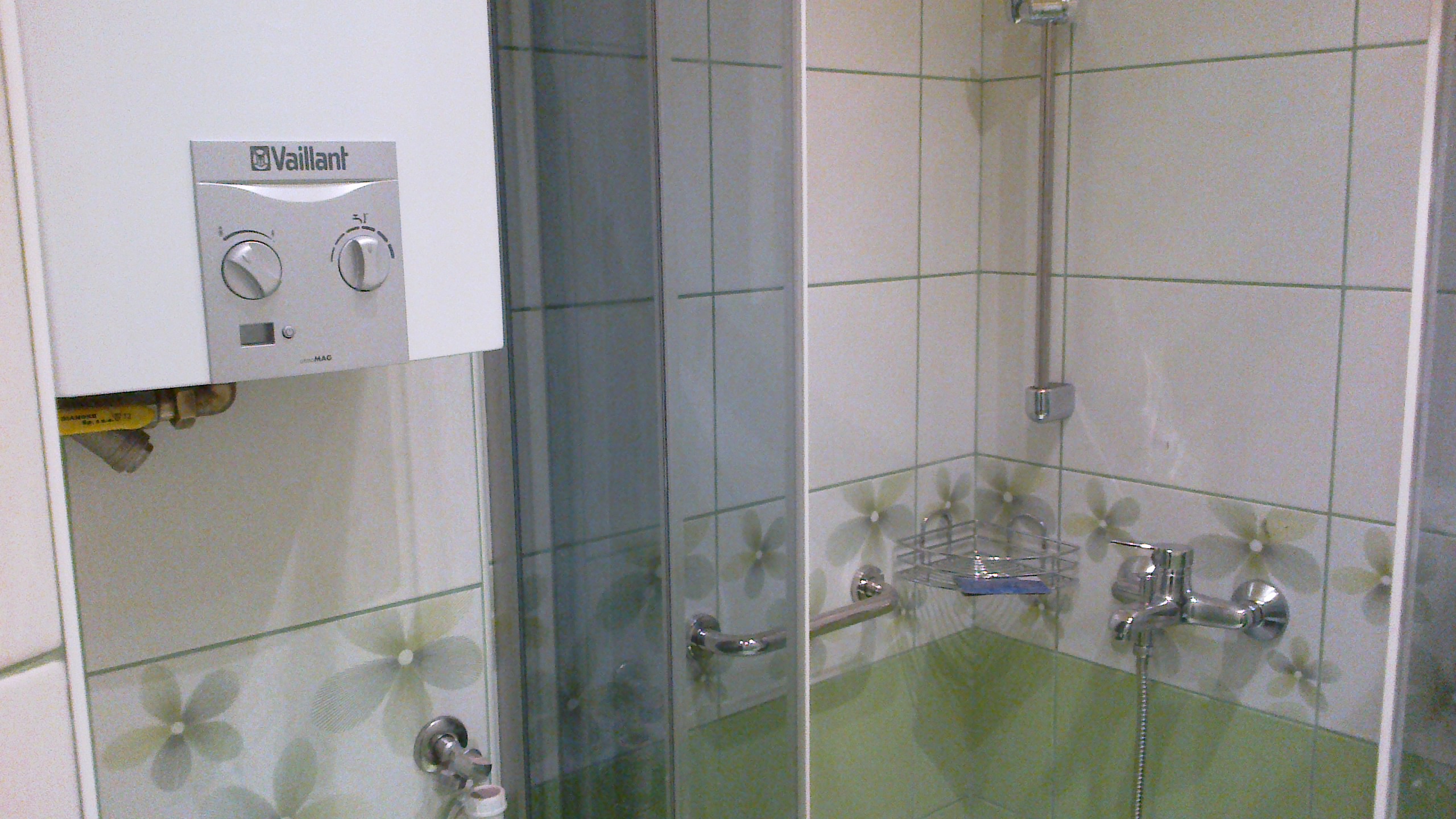 Biały piec gazowy marki Vaillant zamontowany nad umywalką w łazience z kabiną prysznicową, wykończoną kafelkami w biało-zielone kwiaty.