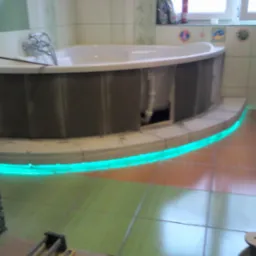 Narożna wanna obudowana płytkami z podświetleniem LED na etapie prac wykończeniowych, widoczne narzędzia i niedokończona podłoga z płytek w dwóch odcieniach zieleni i brązu.