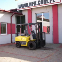 Żółty wózek widłowy GC.POWER FG/FD 15-35 z czarnym masztem, zaparkowany na kostce brukowej przed budynkiem z czerwonymi elementami, widoczny stożek ostrzegawczy.