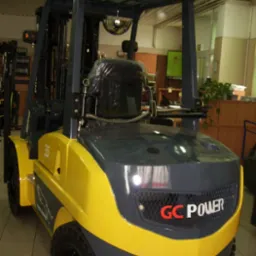 Nowy wózek widłowy GC.POWER FG/FD 15-35 w kolorze żółto-szarym, z czarnym siedziskiem zabezpieczonym folią, prezentowany w hali z widocznymi elementami wyposażenia biurowego w tle.