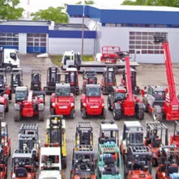 Szeroki kadr na plac z ekspozycją wózków widłowych i ładowarek teleskopowych różnych typów, z budynkiem firmy JMP w tle. Różnorodność modeli i kolorów maszyn.