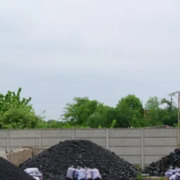 Góra czarnego węgla kamiennego, worki z węglem w dole, betonowy płot w tle, zielone drzewa i pochmurne niebo. Ujęcie z zewnątrz składu.