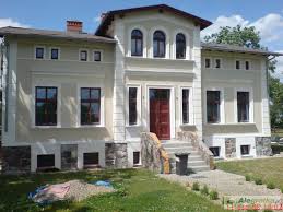 Elewacja odnowionego, dwukondygnacyjnego domu z jasną fasadą, ciemnymi oknami i kamienną podmurówką. Wejście z kamiennymi schodami.