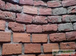 Ceglana ściana z czerwonej i szarej cegły, nierównomierne ułożenie, widoczne spoiny, chropowata faktura, zbliżenie, fragment ściany z logo w rogu.