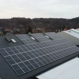 Panele słoneczne zamontowane na dachu budynku z blachy trapezowej, widoczne okna dachowe i fragment krajobrazu z zabudowaniami w tle.