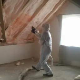 Pracownik w kombinezonie ochronnym aplikuje pianę izolacyjną na skośny dach poddasza, widoczne drewniane elementy konstrukcyjne i przewód doprowadzający pianę.