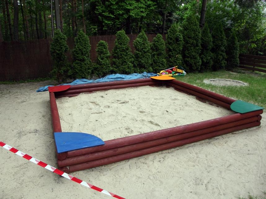 Drewniana piaskownica z kolorowymi narożnikami, wypełniona piaskiem, otoczona zielenią i taśmą ostrzegawczą. Widok z perspektywy.