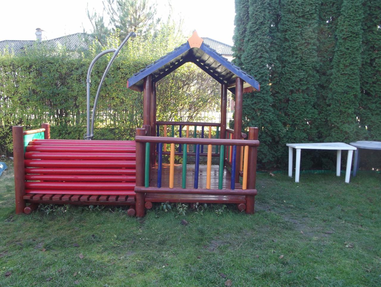 Kolorowy plac zabaw z drewna na trawie, z zadaszeniem i elementami do wspinaczki, otoczony zielenią. Widoczne stoliki i krzesła.