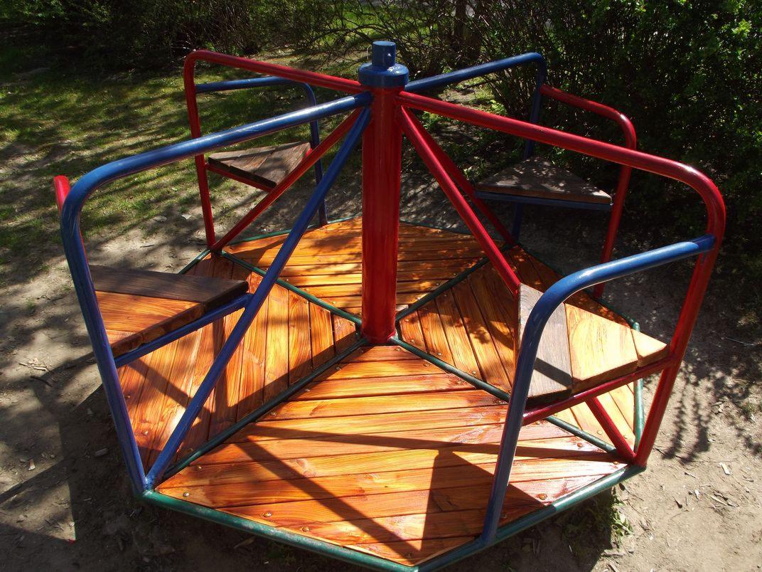 Kolorowa karuzela z drewnianymi siedziskami na placu zabaw, widziana z boku, z zielenią w tle i refleksami słońca na drewnie.