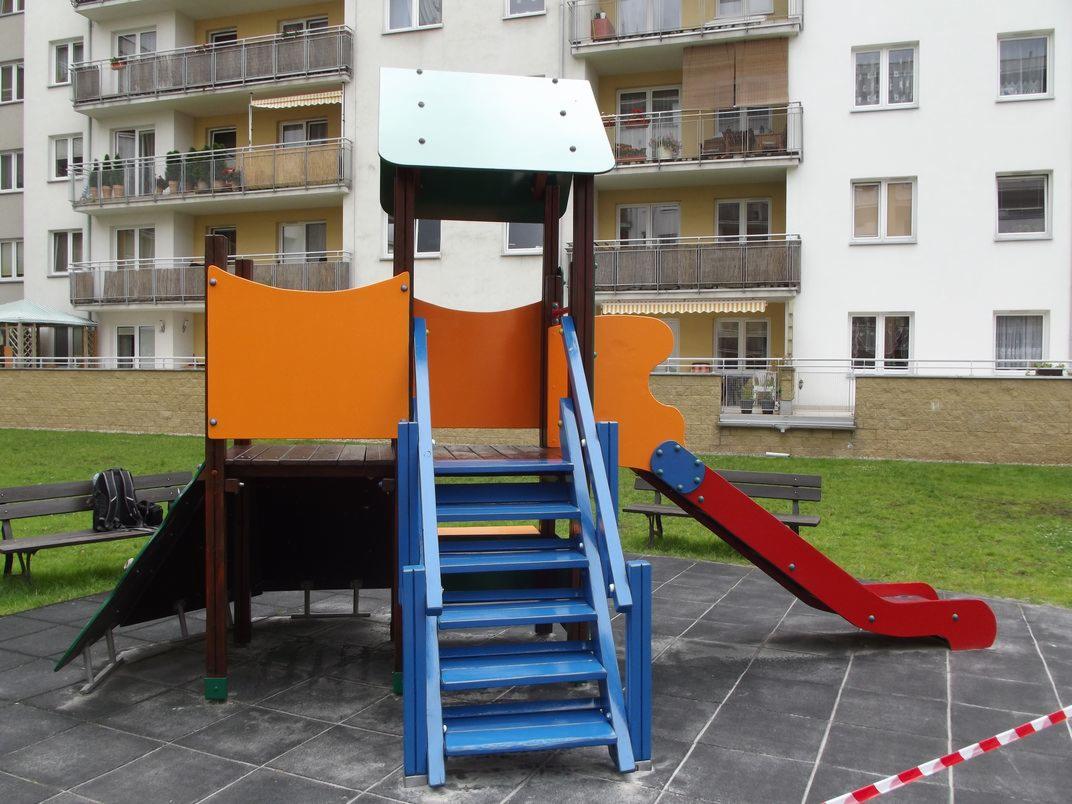 Kolorowy plac zabaw dla dzieci z niebieskimi schodami i czerwoną zjeżdżalnią na tle bloku mieszkalnego z balkonami i zielenią.