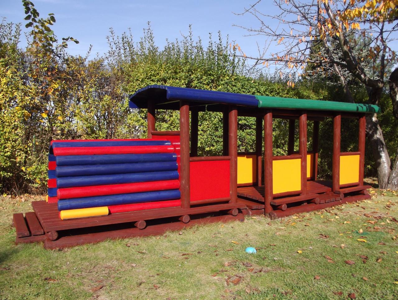 Kolorowy, drewniany pociąg na placu zabaw, z niebiesko-czerwonym wagonem i zielono-żółtymi elementami, otoczony zielenią i drzewami w słoneczny dzień.