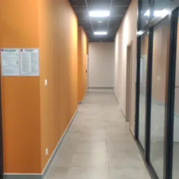 Długi korytarz biurowy z pomarańczowymi ścianami, szarą podłogą z płytek, czarnym sufitem kasetonowym i dużymi przeszkleniami po prawej stronie. Na ścianie po lewej stronie widoczne tablice...