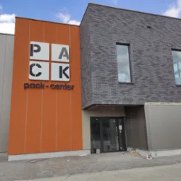 Nowoczesny budynek z pomarańczową elewacją z logo 'PACK-CENTER' i szarą elewacją z cegły, widoczne okna i drzwi wejściowe, kostka brukowa przed budynkiem.
