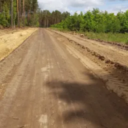 Szeroka droga gruntowa w lesie, świeżo wyrównana, z widocznymi śladami opon ciężkiego sprzętu po bokach i na powierzchni.