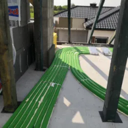 Zielone rury wentylacyjne ułożone na betonowej powierzchni w trakcie montażu, widoczne elementy konstrukcyjne budynku i ocieplenie, w tle dom z ciemnym dachem.