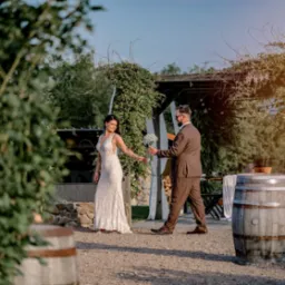 Uśmiechnięta panna młoda w koronkowej sukni odbiera bukiet od pana młodego w brązowym garniturze, w tle pergola porośnięta zielenią i beczki.