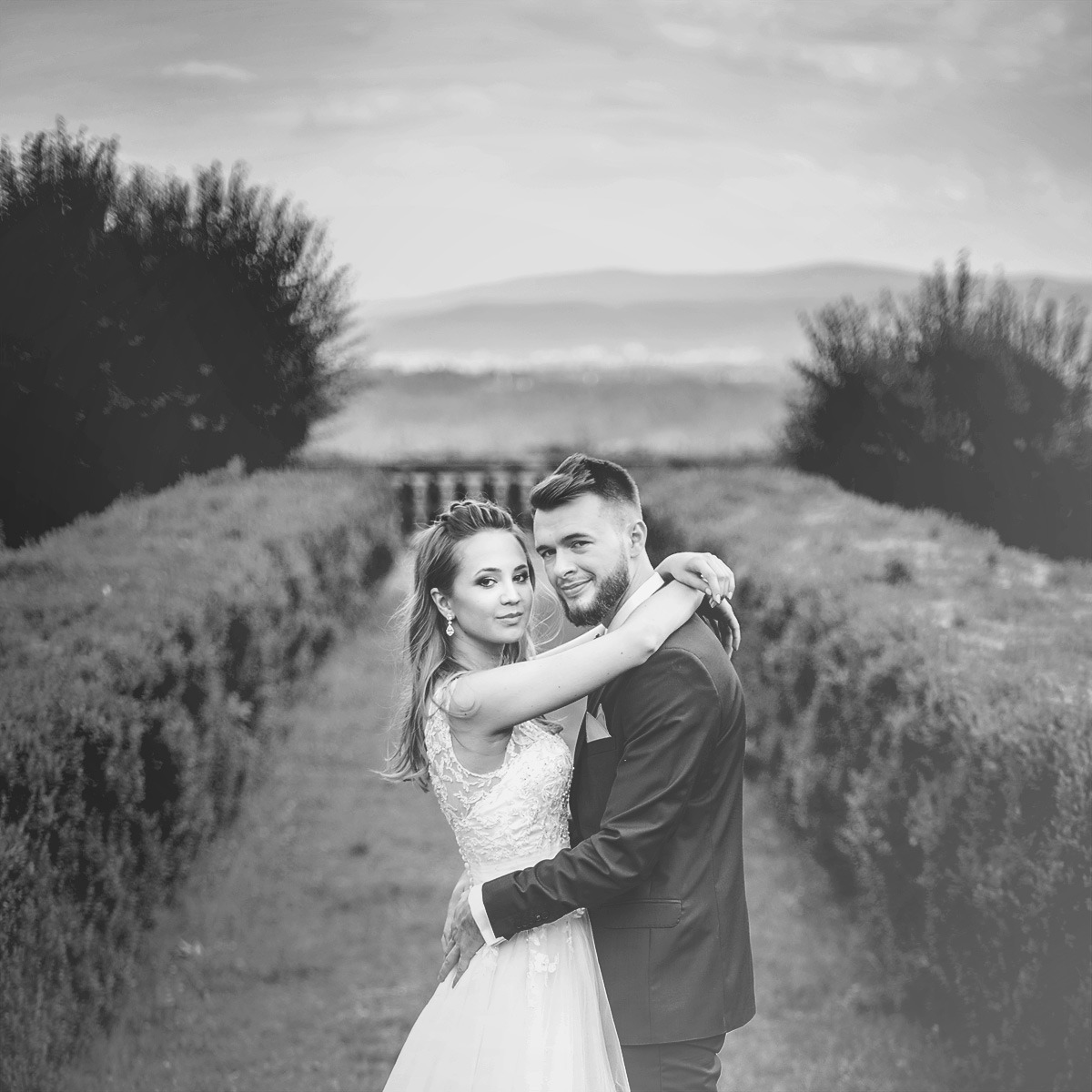 Czarno-białe zdjęcie uśmiechniętej pary młodej w objęciach na tle krajobrazu z bujną zielenią i wzgórzami w oddali.