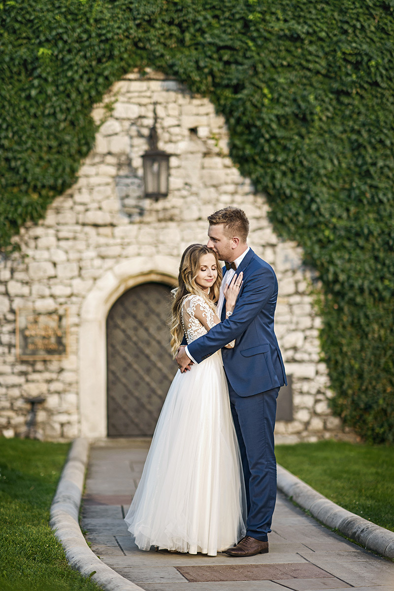 Para młoda w objęciach na tle kamiennej budowli porośniętej bluszczem, panna młoda w białej sukni z koronkowymi rękawami, pan młody w granatowym garniturze.