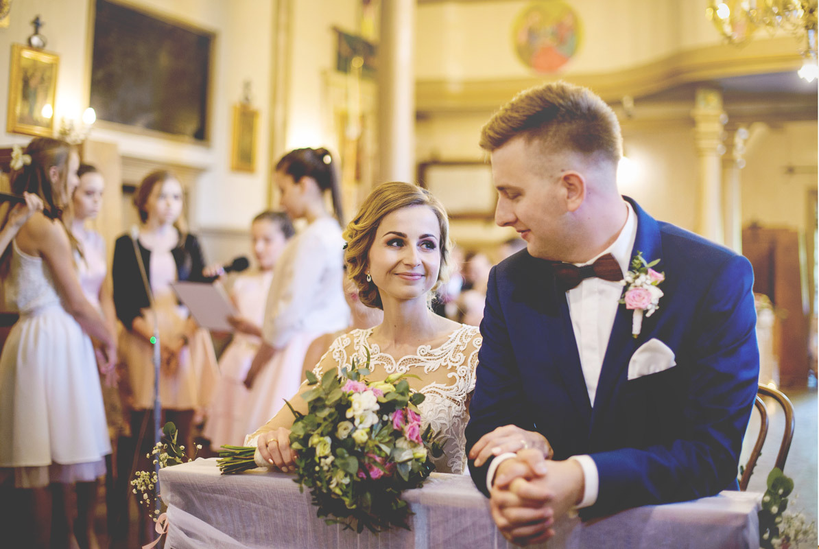 Panna młoda w koronkowej sukni i pan młody w granatowym garniturze z drewnianą muszką podczas ceremonii ślubnej w kościele, w tle druhny.