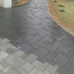 Świeżo ułożony, ciemnoszary chodnik z kostki o nieregularnym wzorze, częściowo mokry, prowadzący od budynku w stronę trawnika z młodymi drzewkami.