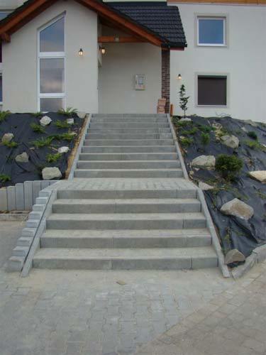 Szerokie, betonowe schody prowadzące do domu z szarą elewacją, z kostki brukowej u podnóża i nasadzeniami roślin na zboczu po bokach.