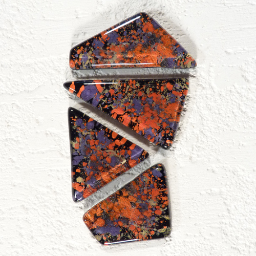 Szklane fragmenty o nieregularnych kształtach z pomarańczowo-fioletową mozaiką na czarnym tle, ułożone w pionową kolumnę na białym, teksturowanym tle.