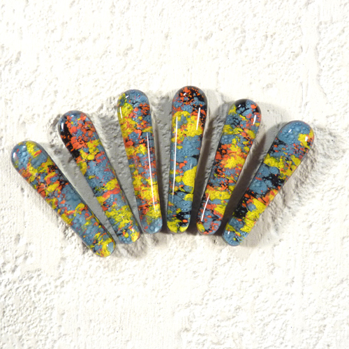 Sześć szklanych elementów biżuterii o podłużnym kształcie, pokrytych wzorem z niebieskich, żółtych, pomarańczowych i czarnych plam na teksturowanym, jasnym tle.