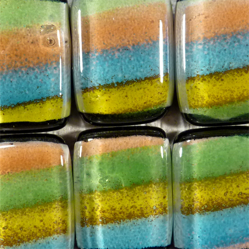 Sześć szklanych, artystycznych kostek z zatopionymi, kolorowymi pasami: żółtym, turkusowym, pomarańczowym i zielonym. Kostki ułożone blisko siebie, tworząc mozaikę z efektem gradientu.