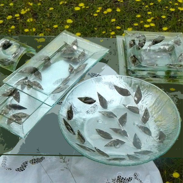 Zestaw szklanych naczyń z motywem liści na tle łąki z żółtymi kwiatami; okrągła misa, prostokątny talerz, kwadratowe talerzyki i mały półmisek.