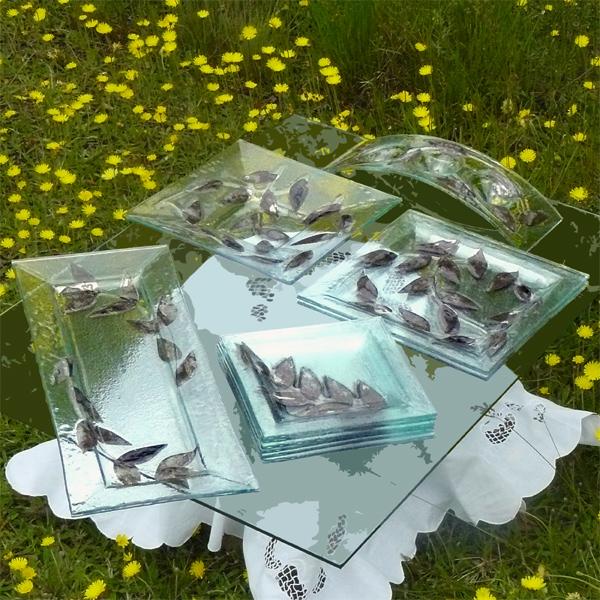 Szklane naczynia dekoracyjne w kształcie liści na szklanym stole z haftowanym obrusem, na tle łąki z żółtymi kwiatami.