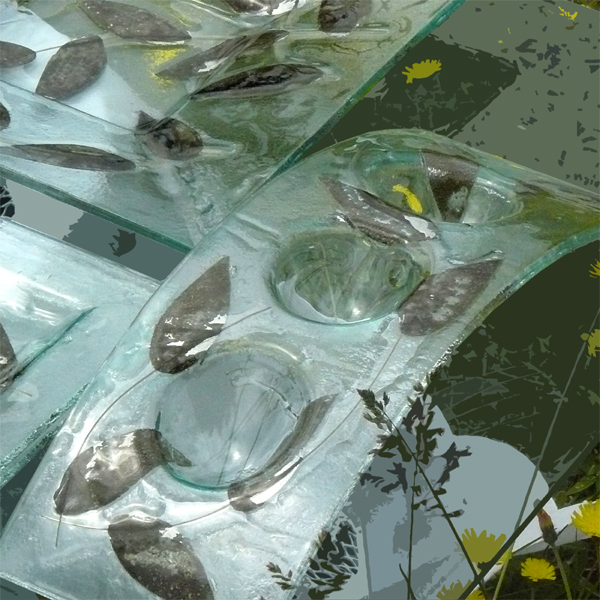 Szklane naczynia dekoracyjne z zatopionymi liśćmi, część zestawu prezentowego, widoczne żółte kwiaty w tle.