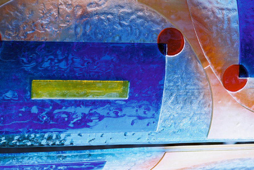 Abstrakcyjna kompozycja z kolorowego, fakturowanego szkła artystycznego: niebieskie i żółte prostokąty, czerwone koła z wyciętymi ćwiartkami, na tle gradientu od błękitu do pomarańczy.