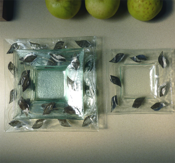 Dwa szklane kwadratowe naczynia z motywem liści, większe i mniejsze, ułożone obok siebie na blacie, z trzema zielonymi jabłkami w tle.