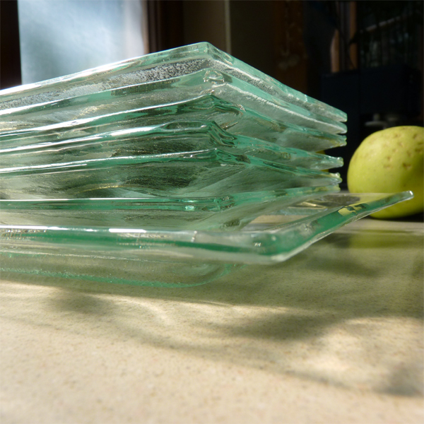 Stos szklanych, kwadratowych talerzy o nieregularnej fakturze, delikatnie zielonkawych, ułożonych na blacie, z zieloną gruszką w tle.