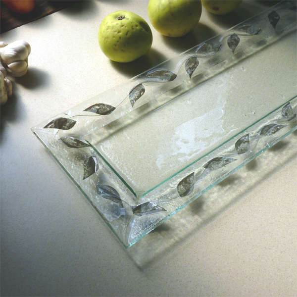 Prostokątna, szklana taca dekoracyjna z motywem liści na obrzeżach, obok jabłek i czosnku na jasnym blacie.