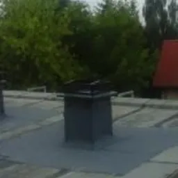 Szary, płaski dach z papy z kominem w centralnej części kadru, w tle zielone drzewa i fragment czerwonego dachu sąsiedniego budynku.