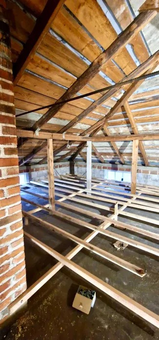 Konstrukcja drewniana poddasza z widocznymi elementami więźby dachowej i legarami podłogowymi, ceglany komin w rogu, okno w tle. Prace remontowe w surowym stanie.