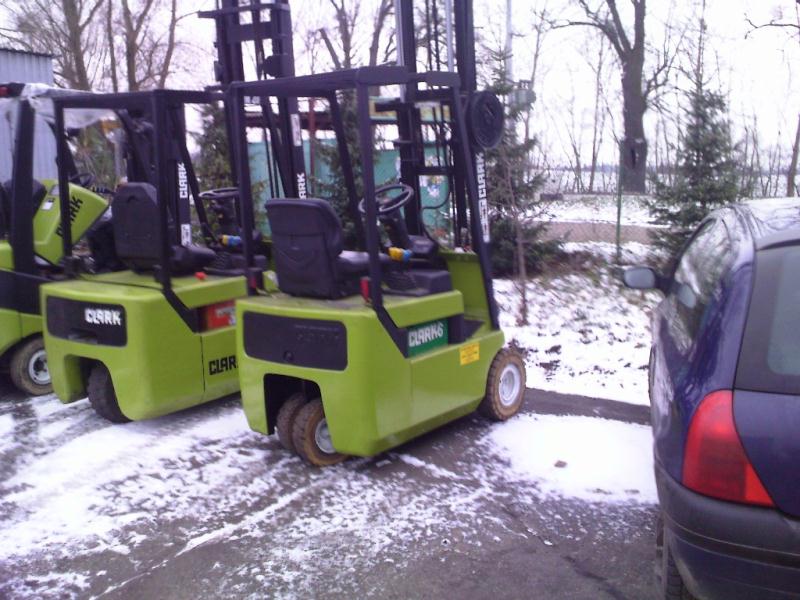 Rząd nowych, zielonych wózków widłowych marki CLARK CTM 12s, zaparkowanych na placu pokrytym śniegiem, z widocznym fragmentem niebieskiego samochodu po prawej stronie kadru i drzewami w tle.