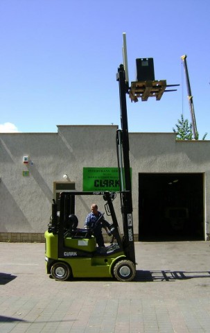 Zielony wózek widłowy Clark CGP 18 podnosi paletę z ładunkiem na tle budynku z zielonym szyldem reklamowym.