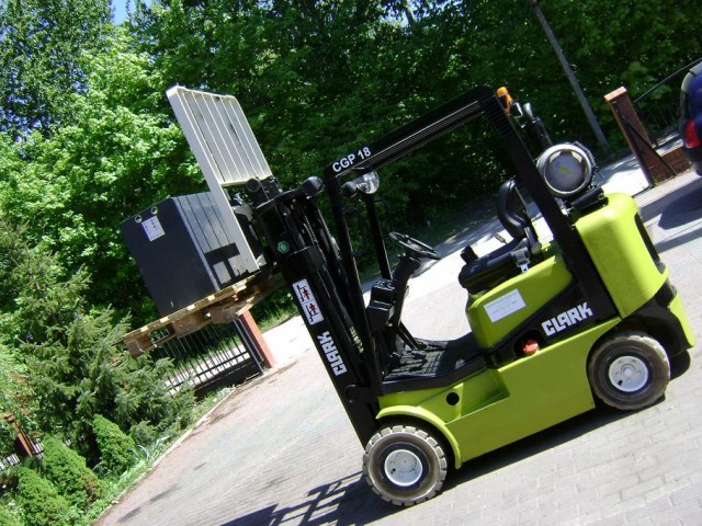 Żółto-czarny wózek widłowy Clark CGP 18 na zewnątrz, podnosi paletę z czarnym ładunkiem, w tle zieleń i brama.