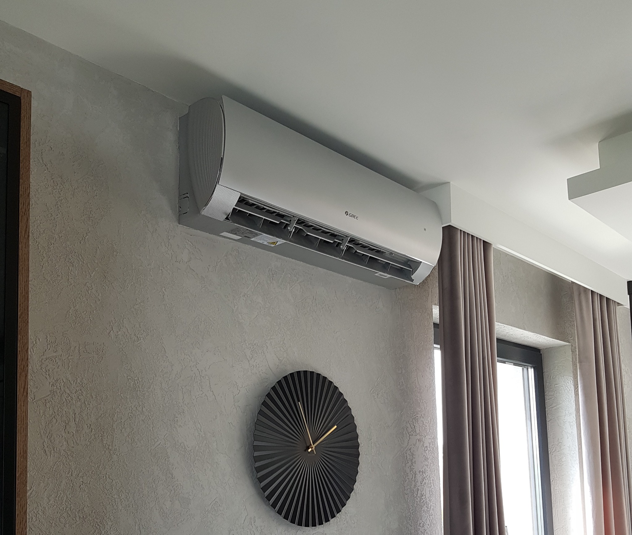 Nowoczesny klimatyzator marki Gree zamontowany na ścianie o fakturze betonu architektonicznego, poniżej dekoracyjny zegar w stylu art deco, w tle zasłonięte okno.