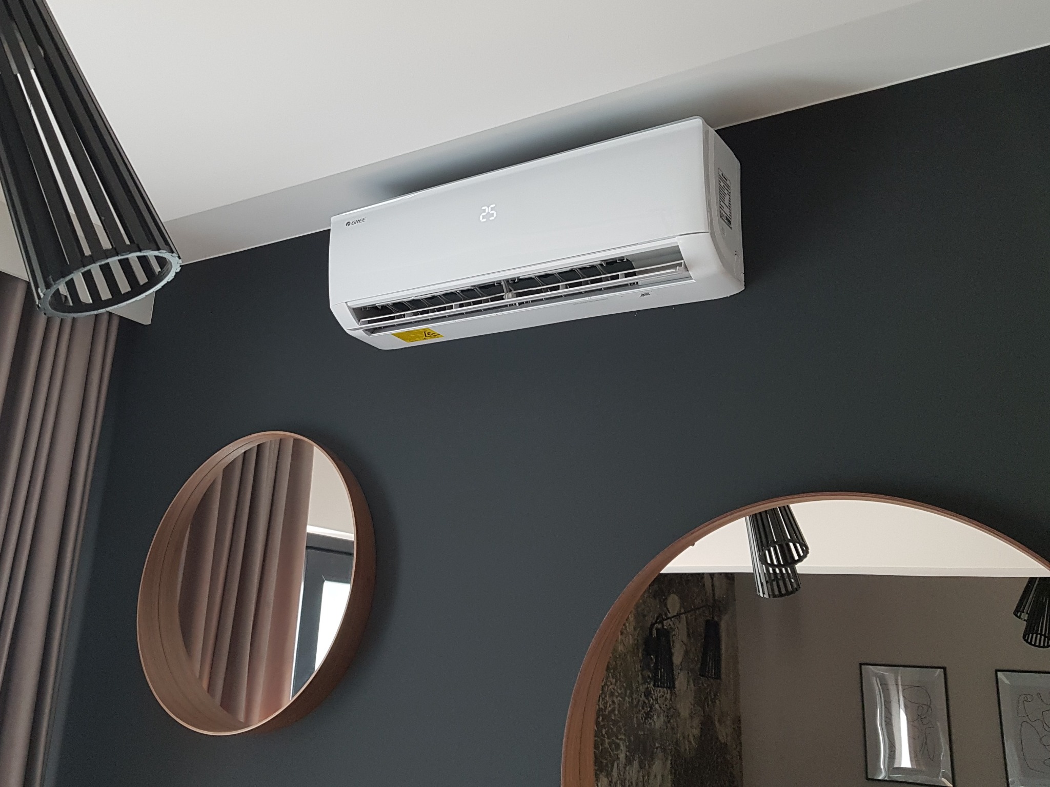 Biały klimatyzator marki Gree zamontowany na ciemnoszarej ścianie w salonie, widoczne dwa okrągłe lustra z drewnianą ramą oraz lampa z czarnymi kloszami.