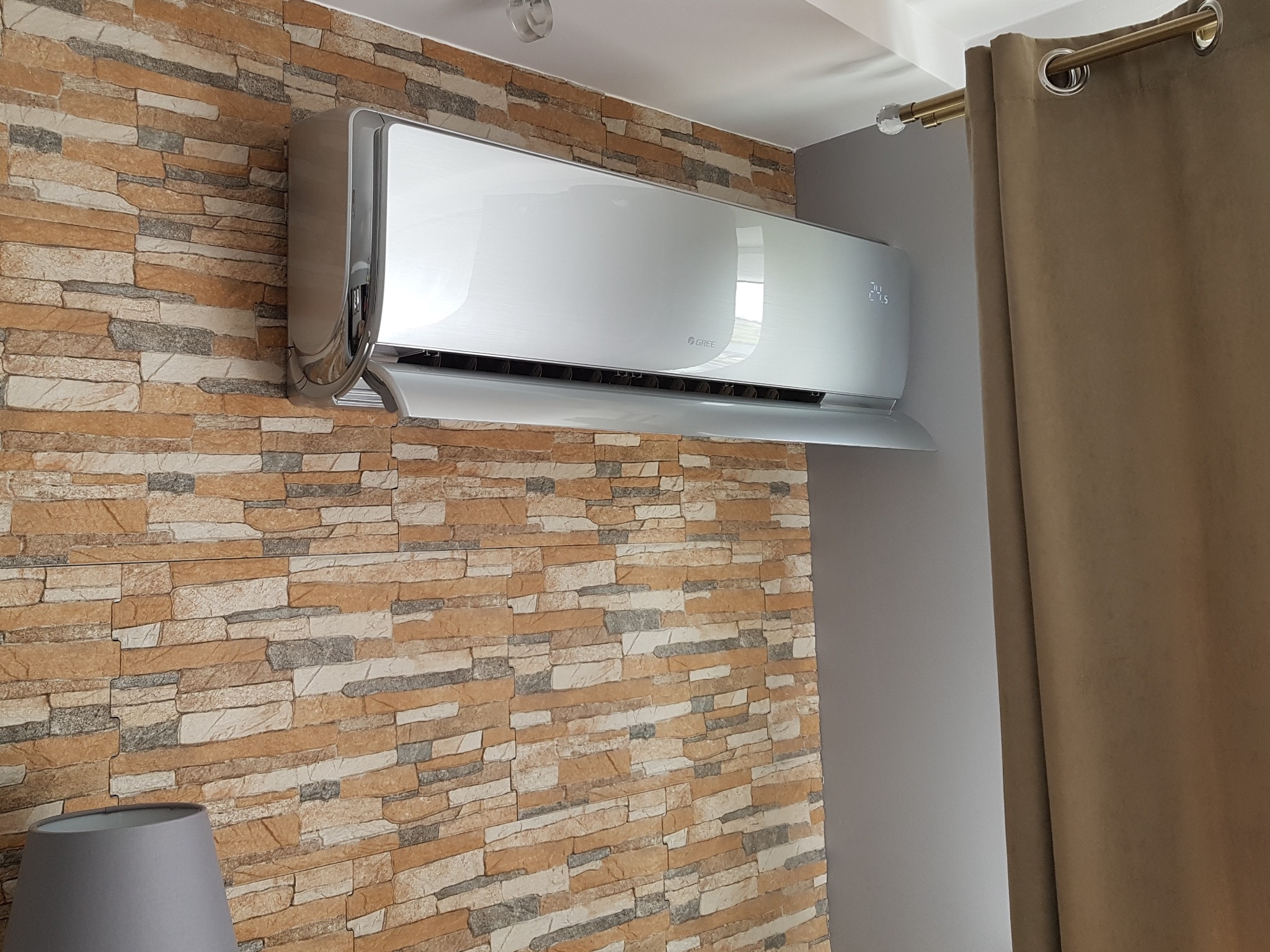 Nowoczesny klimatyzator marki Gree zamontowany na ścianie z dekoracyjnej cegły, obok zasłony w odcieniach brązu.