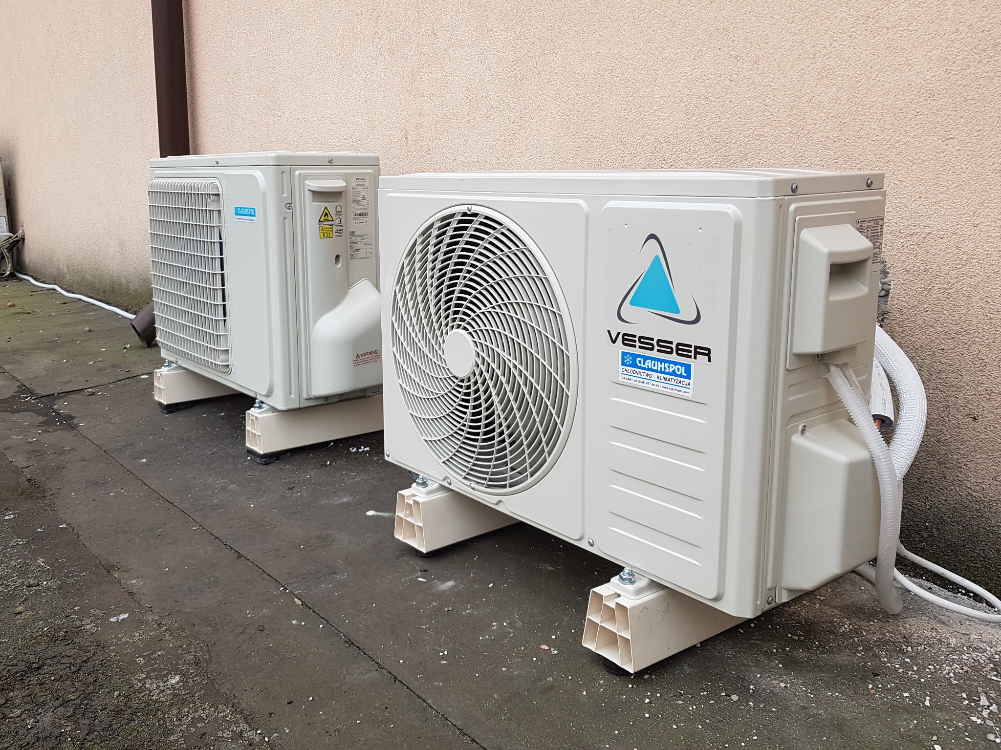 Dwa zewnętrzne agregaty klimatyzacyjne marki Vesser CLAUHSPOL zamontowane na betonowym podłożu przy ścianie budynku. Widoczne białe rury odprowadzające skropliny.