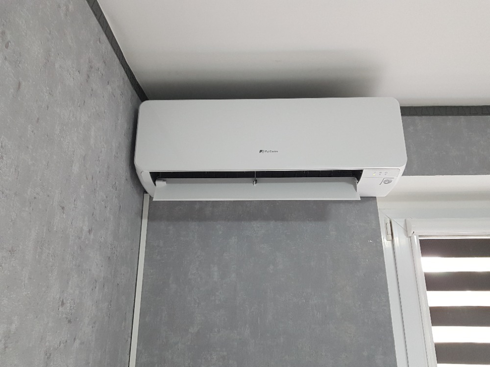 Biały klimatyzator zamontowany na szarej ścianie o teksturze betonu, widoczne okno z żaluzjami w tle.