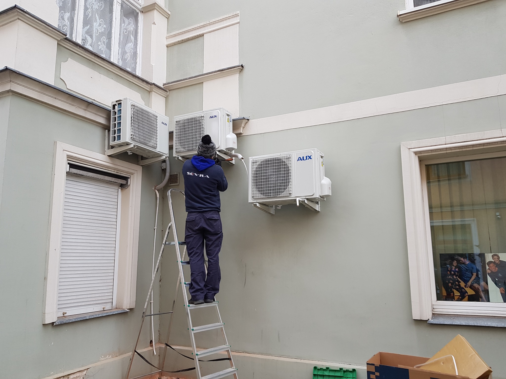 Instalator na drabinie montuje trzy klimatyzatory AUX na elewacji budynku z oknami ze szprosami i roletami zewnętrznymi.